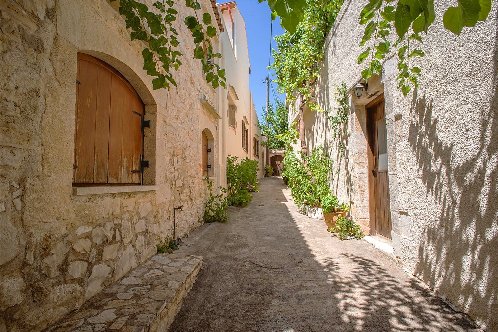 A narrow lane in Douliana between old stone houses, with arched doorways, potted plants and vines overhead