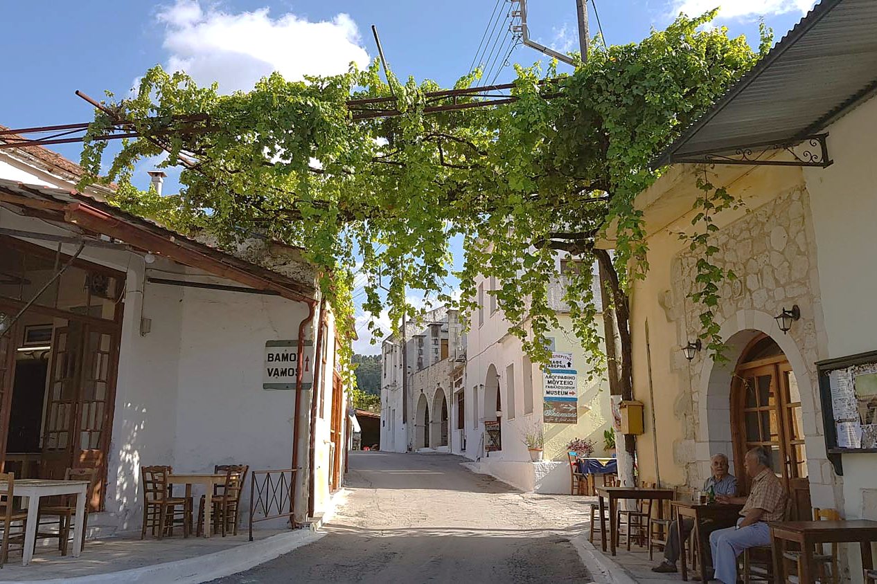 The main street of Gavalochori shaded by a vine pergola, with taverna tables set out beneath it