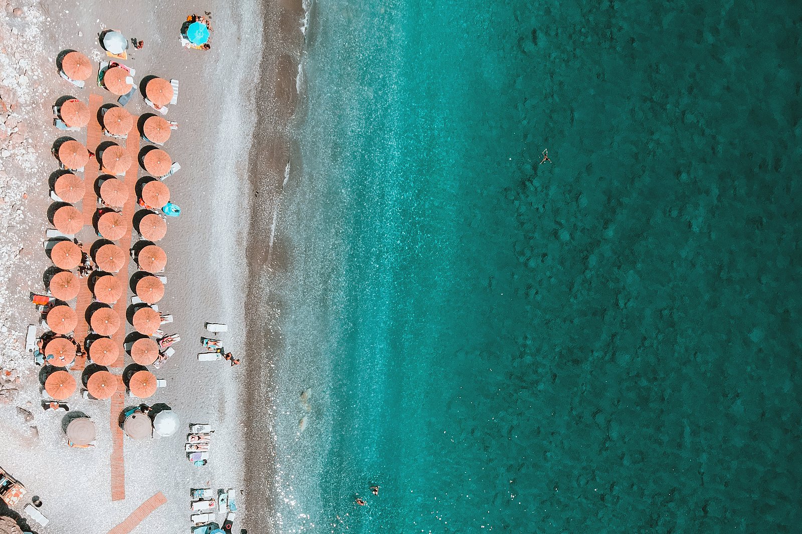 Rows of orange parasols on a beach beside clear turquoise water, seen from above