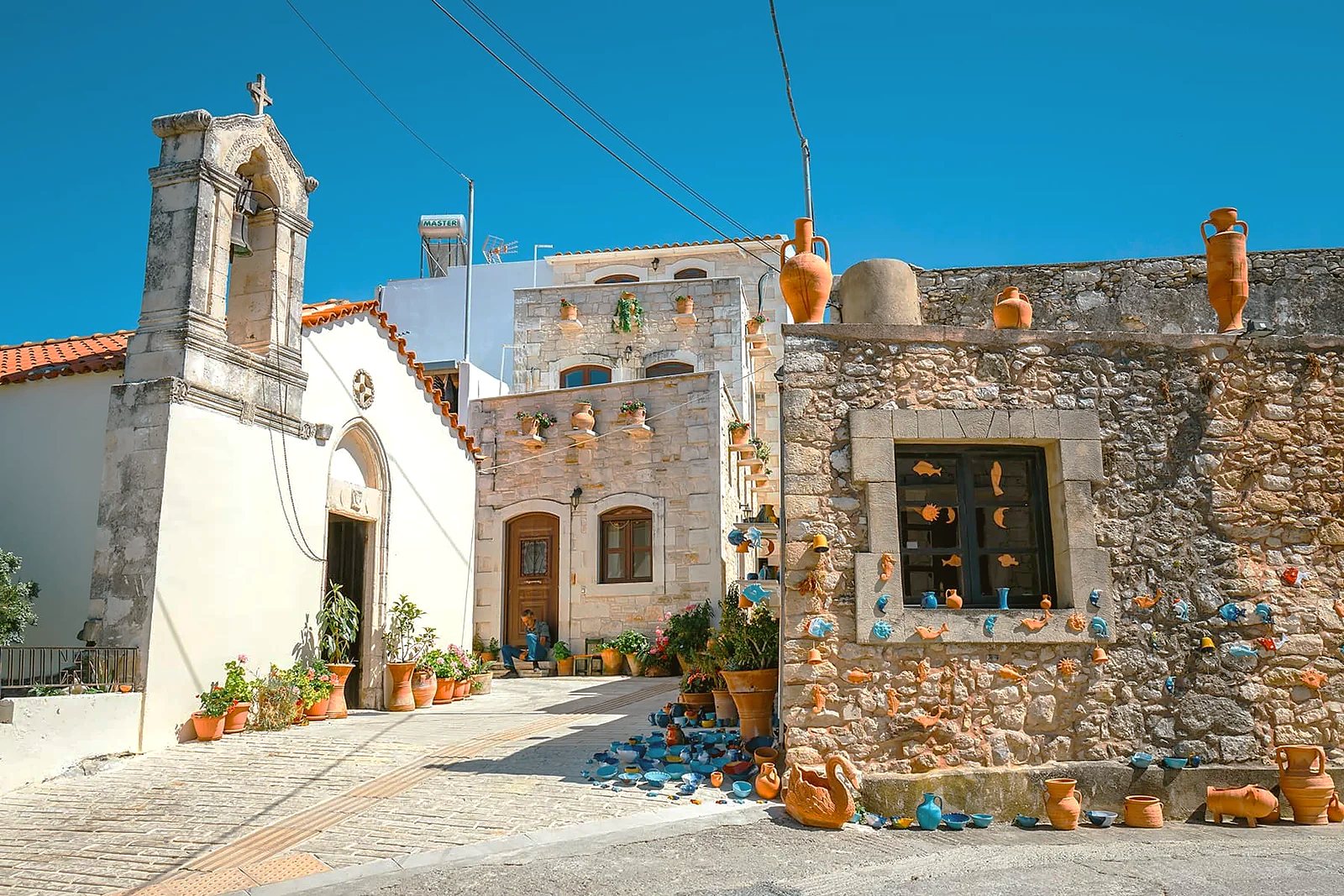 The square at Margarites, with a stone belfry and terracotta pots and blue glazed ceramics set out along a workshop wall