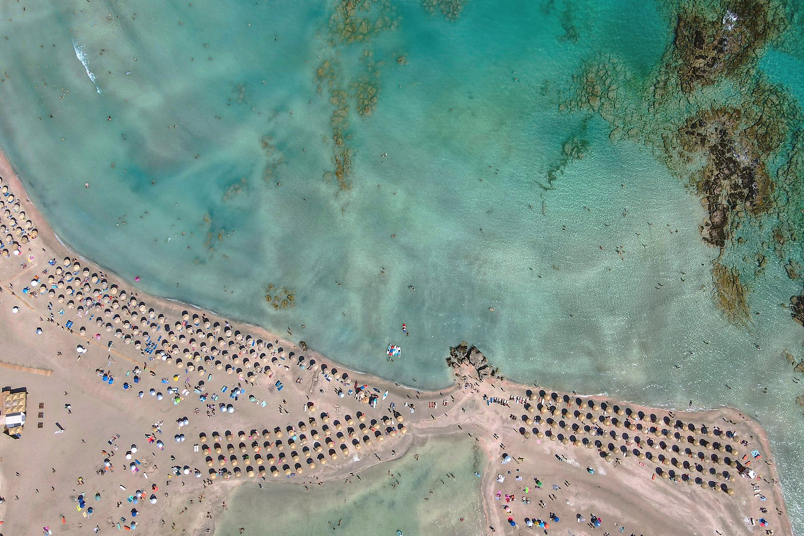 The shallow lagoon at Elafonisi from the air, with rows of umbrellas along the pale sandbar and clear turquoise water either side