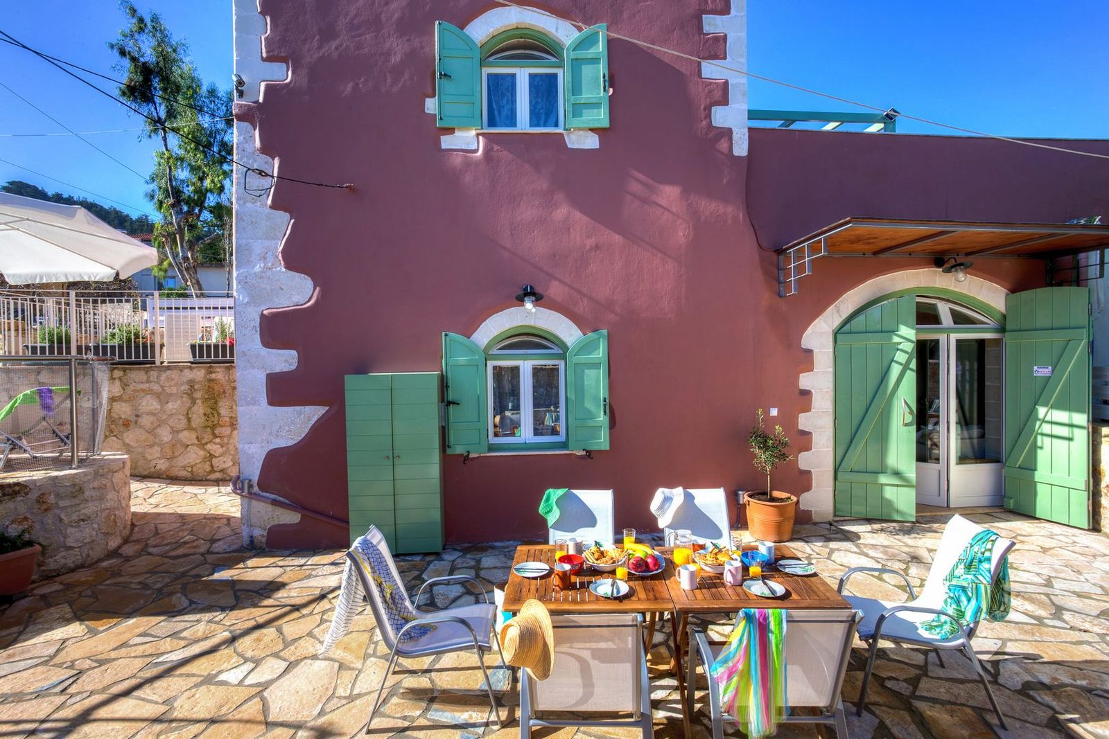 The plum-coloured facade of Horizon Villa Likotinara with green shutters and a stone terrace laid for lunch